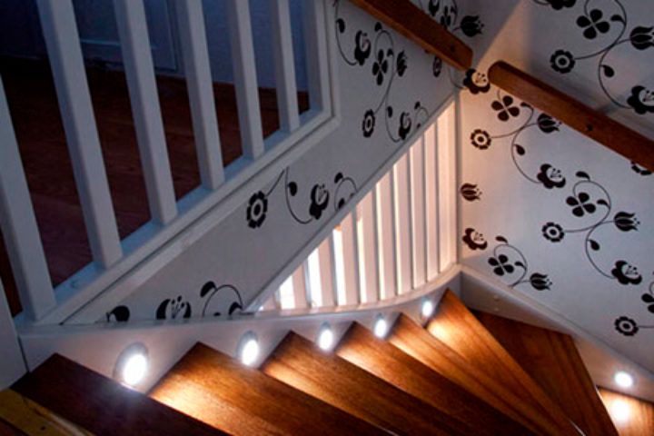 Spot lys ned en trapp med tre gelender og trappetrinn med hvitt tapet på veggene som har sort blomstermønster på seg