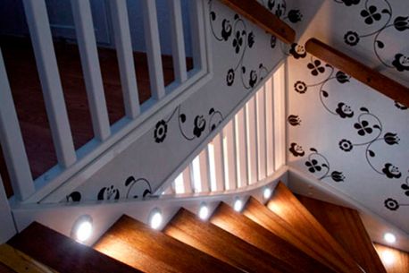 Spot lys ned en trapp med tre gelender og trappetrinn med hvitt tapet på veggene som har sort blomstermønster på seg