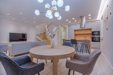 Lyst interiør med åpen planløsning til et hvitt kjøkken, et rundt spisebord med grå stoler rundt, en grå sofa foran en tv som er godt opplyst av en stor lampe over spisebordet og spotlys i det hvite taket