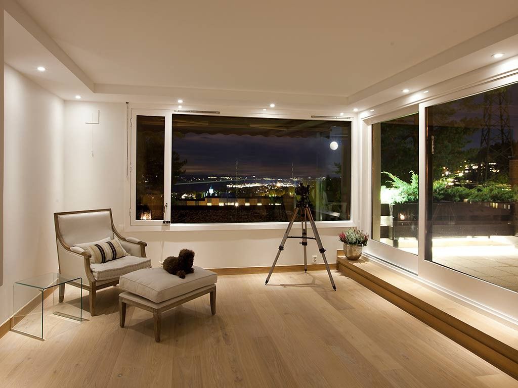 Et lyst rom med hvite vegger og en hvit lenestol med spotlys i taket som har en hevet del. Store vindu som viser utsikten på kveldstid